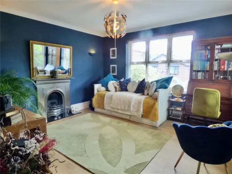 The living room features dark blue walls and a decorative fireplace, with a large mirror reflecting elegance. A daybed adorned with pillows rests on a green rug beside a chair. A wooden cabinet with shelves adds charm, while the large window allows natural light to job template its way in.